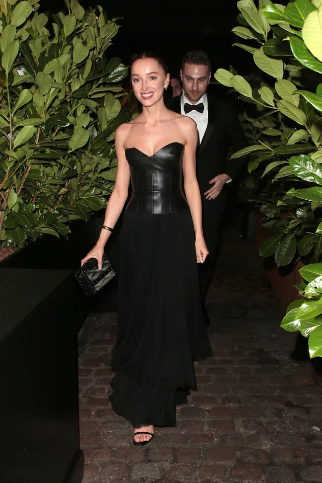 Phoebe Dynevor walking beside foliage in a black dress and heeled sandals.