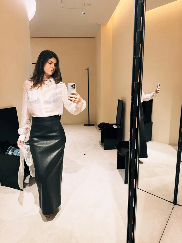Alexandra Daddario taking a mirror portrait in a white blouse and long dark skirt.