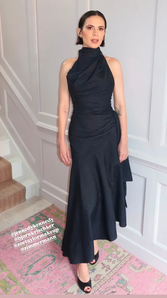 Hayley Atwell standing indoors in a black halter dress.