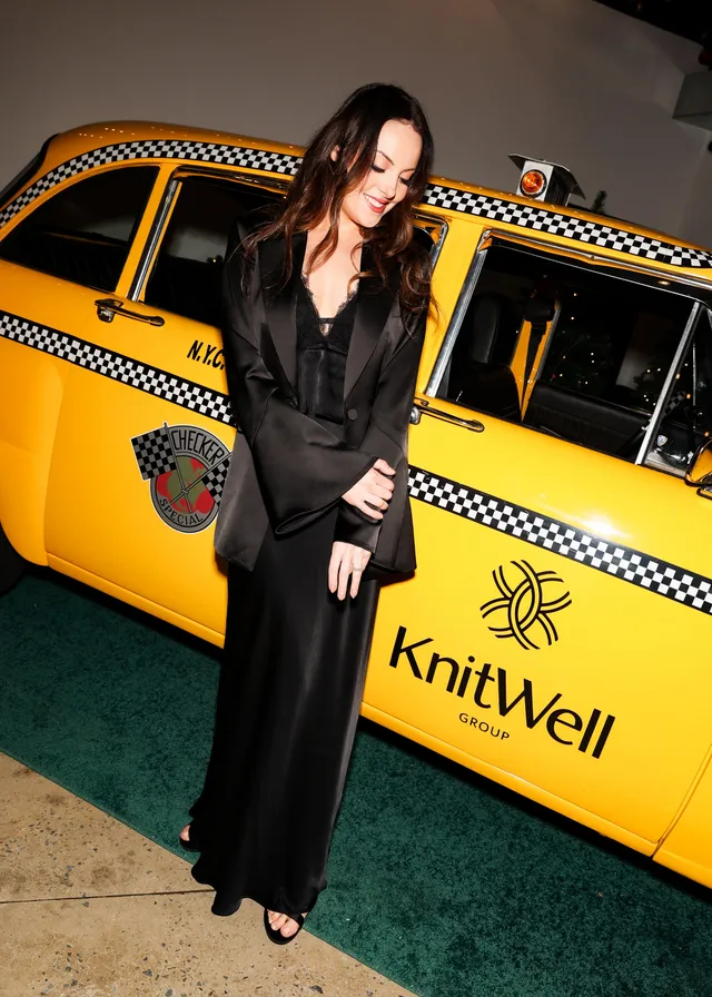 Elizabeth Gillies standing beside a yellow taxi in a long black outfit.