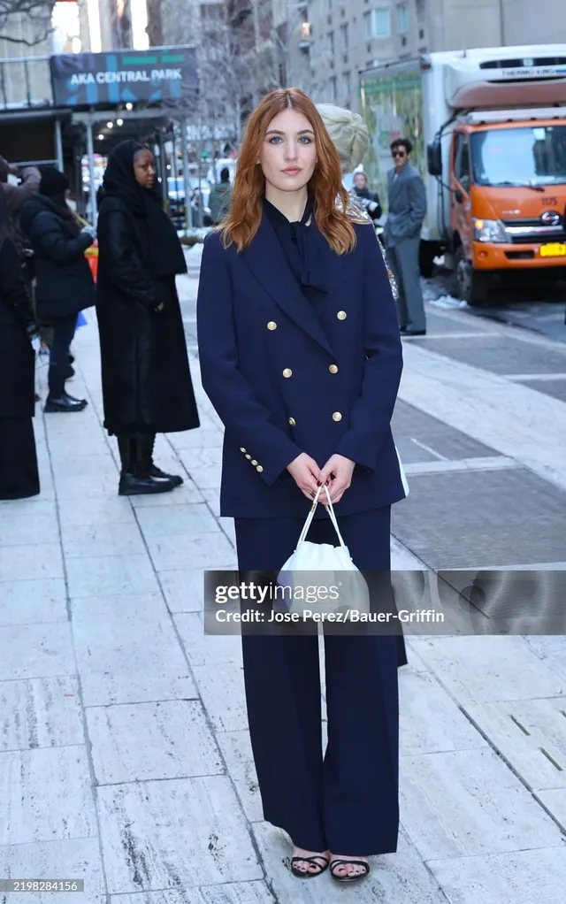 Sophie Nélisse standing outside in a dark trouser suit and heeled sandals.