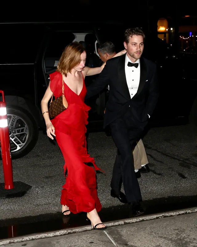 Phoebe Dynevor arriving at an event in a red dress beside a companion.