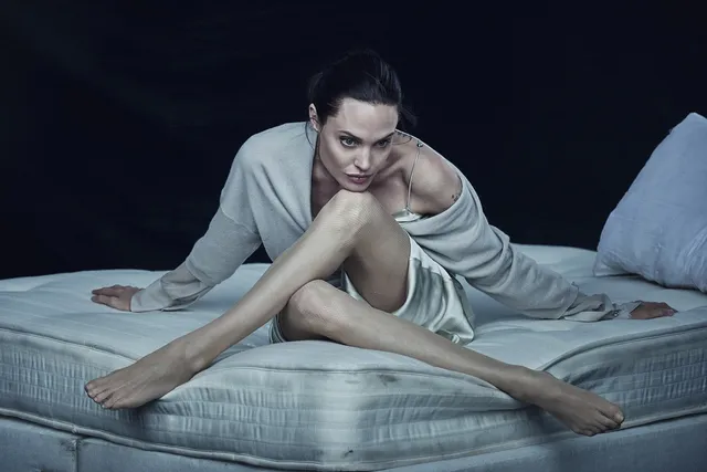 Angelina Jolie seated barefoot on a bed in a pale robe.