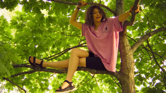 Odessa A'zion sitting in a tree wearing a pink shirt and sandals.