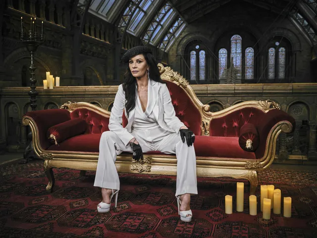 Catherine Zeta-Jones seated on a red sofa in a white trouser suit.