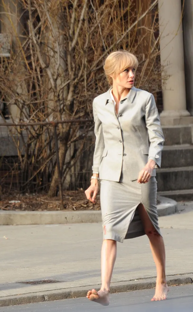 Angelina Jolie standing barefoot outside in a gray skirt suit.