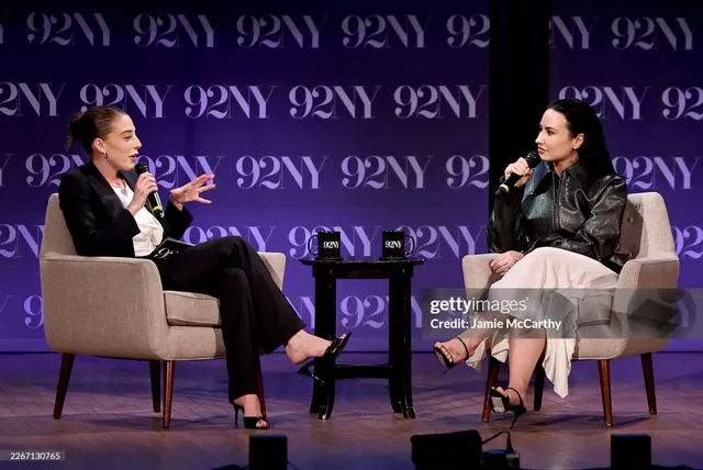 Demi Lovato sitting on stage during an interview in a skirt and heels.