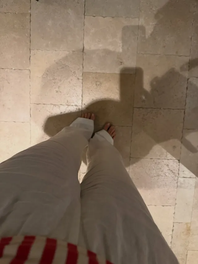 Hailee Steinfeld standing barefoot on a tiled floor in loose trousers.