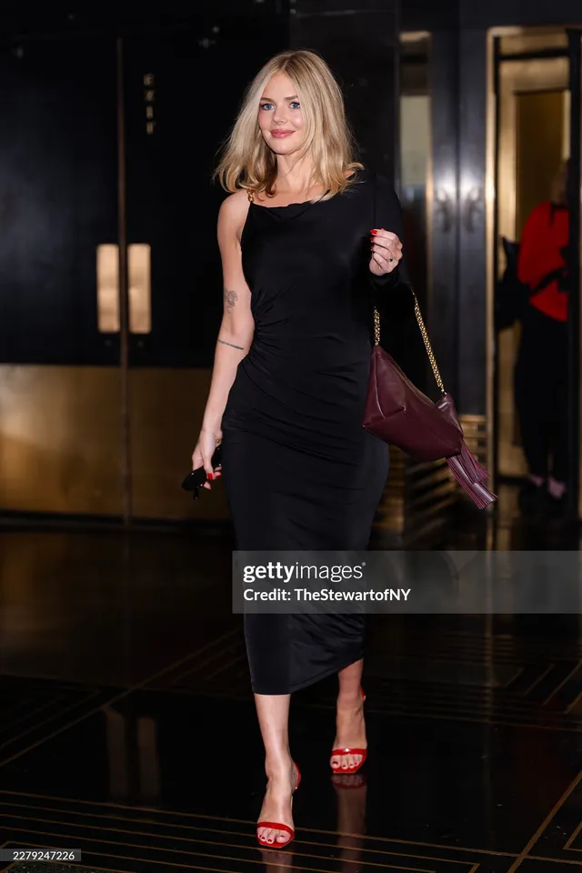 Samara Weaving walking indoors in a black dress and red heels.