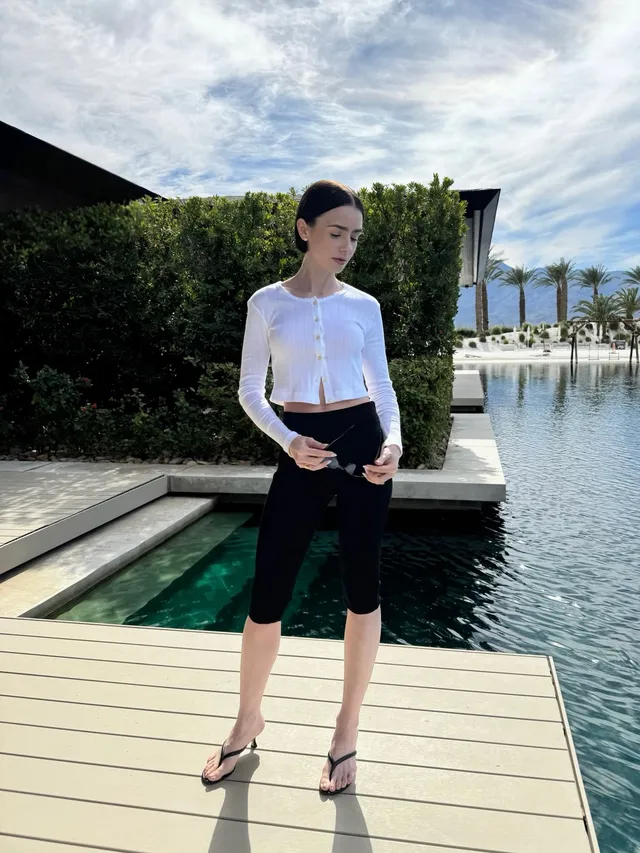 Lily Collins standing by water in cropped trousers and flip-flops.