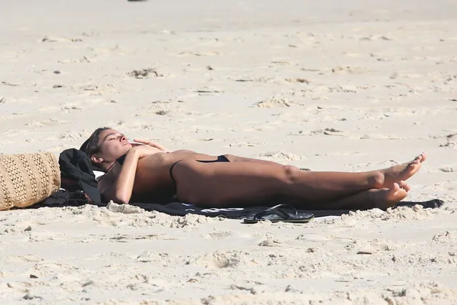 Bruna Marquezine reclining barefoot on sand in a dark bikini.
