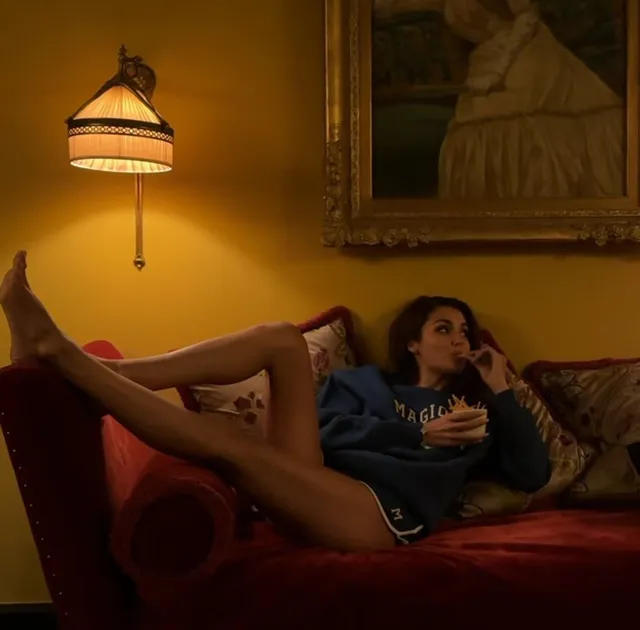 Hande Erçel reclining barefoot on a red sofa in a T-shirt and shorts.