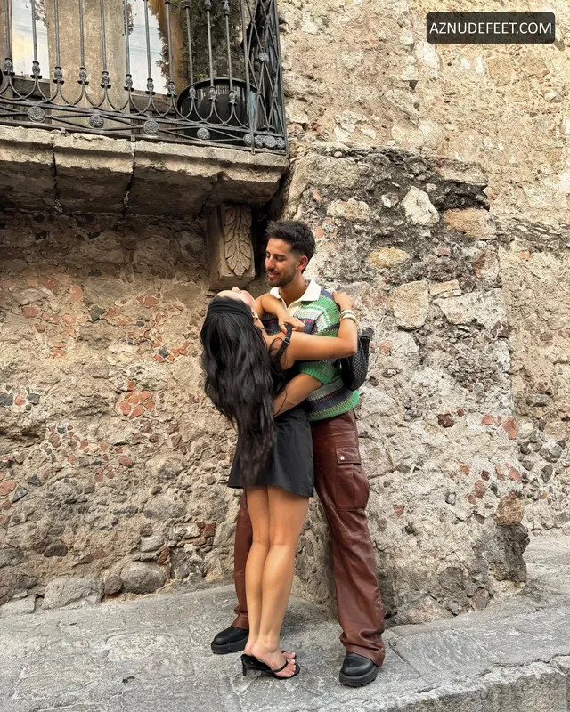 Cierra Ramirez in a dark casual outfit and flat sandals beside her partner on a stone street.