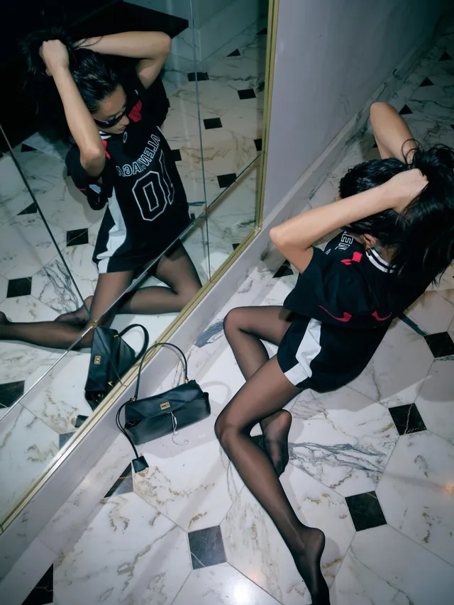 Shay Mitchell posing in front of a mirror in a dark outfit and heels.