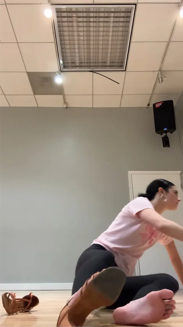 Charli D’Amelio sitting on a rehearsal-room floor with bare soles visible.