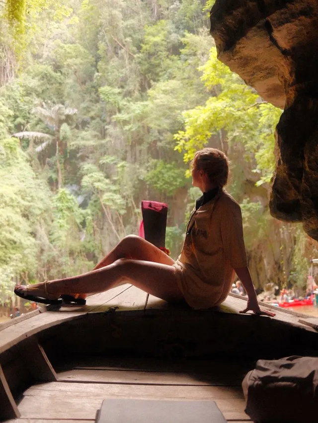 Kathryn Newton sitting beside a cave opening in a pale outfit and dark heels.