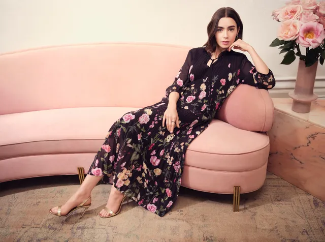 Lily Collins sitting barefoot on a pink sofa in a flowered dress.