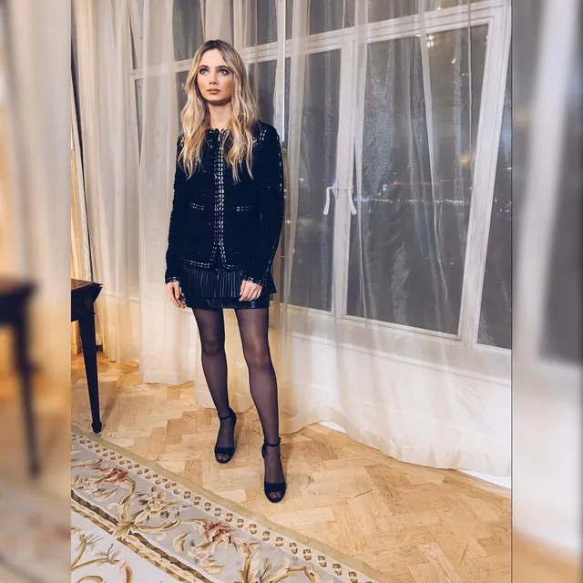 Freya Allan posing indoors in a dark outfit, tights and black heels.