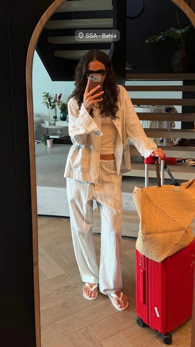 Bruna Marquezine taking a mirror selfie beside luggage in a pale outfit and flat sandals.