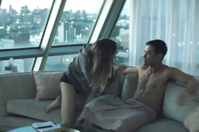 Halston Sage sitting barefoot beside a man on a sofa in a scene.