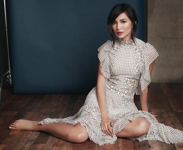 Gemma Chan sitting on a wooden floor in a patterned dress.