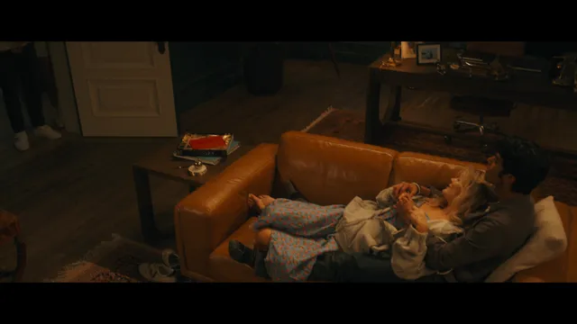 Emily Alyn Lind reclining barefoot on a sofa in a film scene.