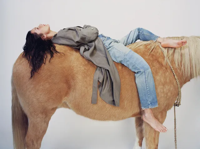 Bella Hadid reclining across a horse in jeans with her bare feet extended.