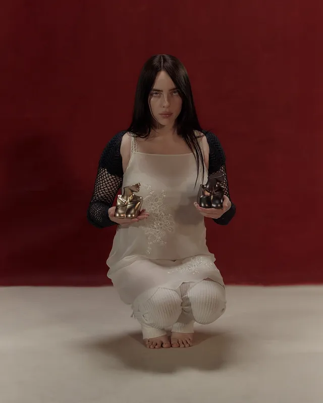 Billie Eilish a kneeling cream outfit against a red backdrop.