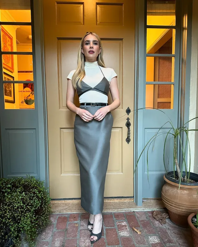 Emma Roberts standing outside a doorway in a grey dress and heels.