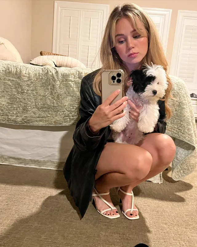 Brec Bassinger crouching in pale heels while holding a dog for a mirror selfie.