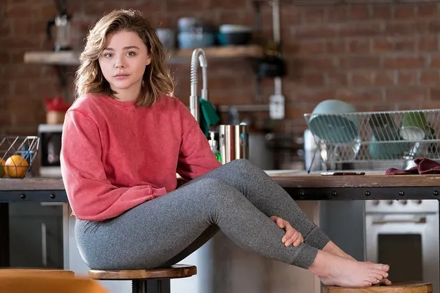 Chloë Grace Moretz sitting barefoot on a kitchen counter in a red sweater.
