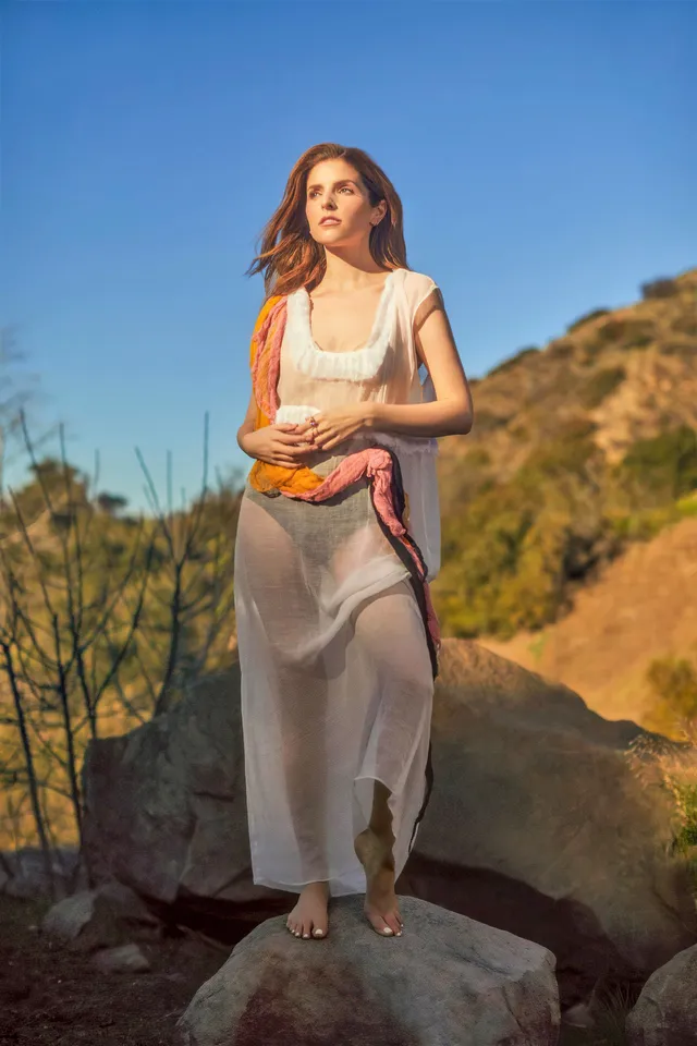 Anna Kendrick standing barefoot on rocks wearing a pale dress.