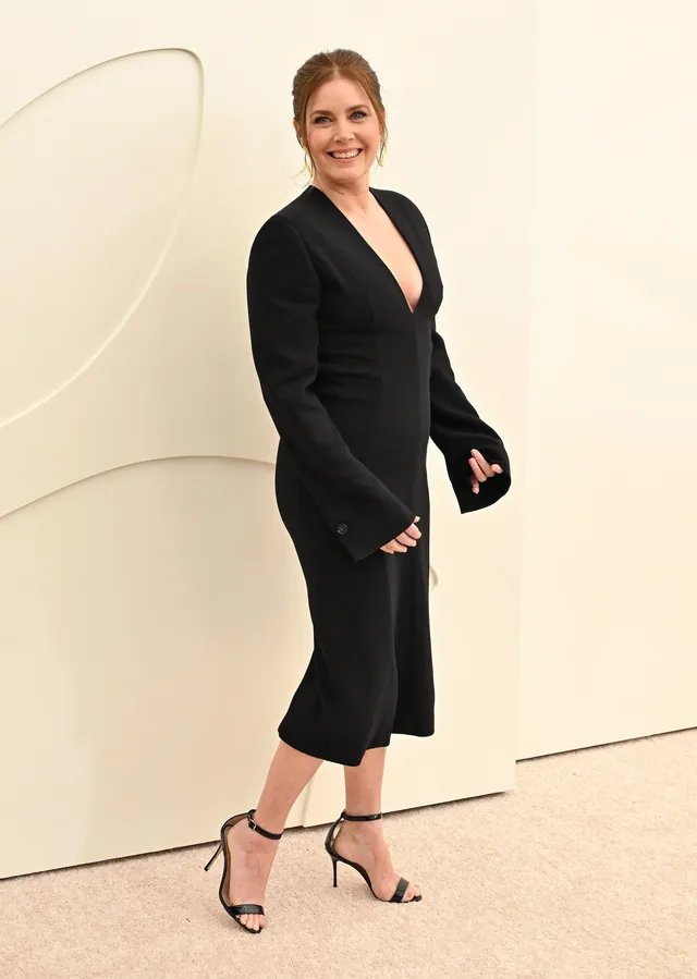 Amy Adams walking indoors in a black dress and sandals.