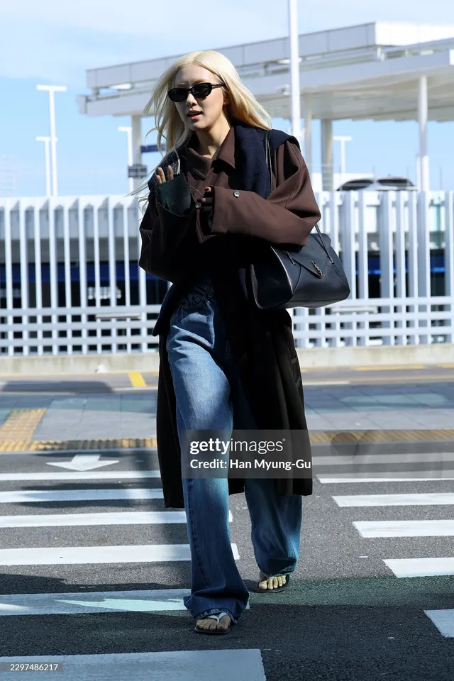 Rosé walking outside in a long brown coat, blue jeans and dark sandals.
