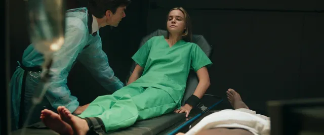 Bailee Madison reclining barefoot in hospital clothing in a scene.