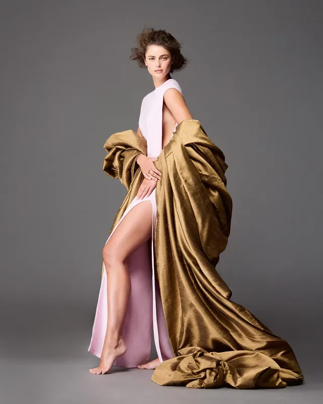 Taylor Hill standing barefoot in a studio wearing a pale pink dress and a long coat.