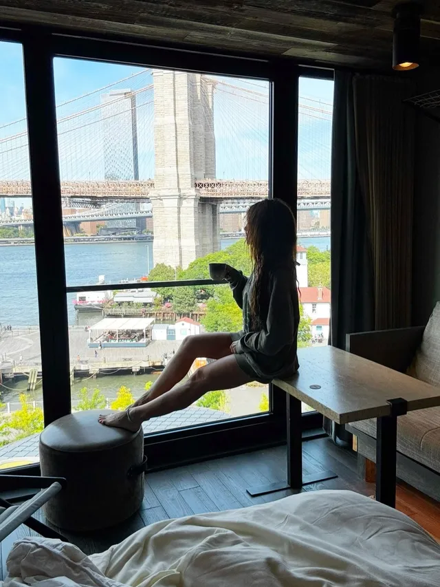 Sarah Shahi sitting beside a window overlooking a bridge.