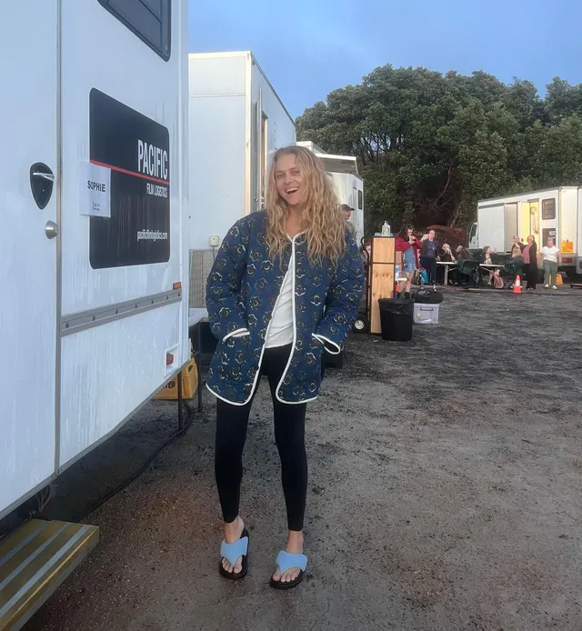 Teresa Palmer standing beside a trailer in dark trousers and blue shoes.