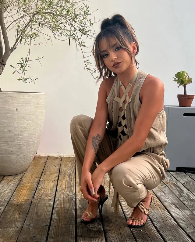 Isabela Merced crouching outdoors in a pale outfit and strappy heels.
