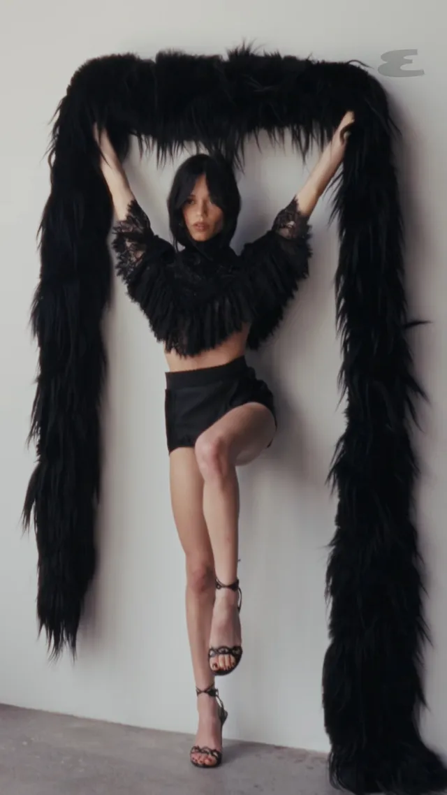 Jenna Ortega posing in a black cropped outfit with feathered fabric and strappy heels.
