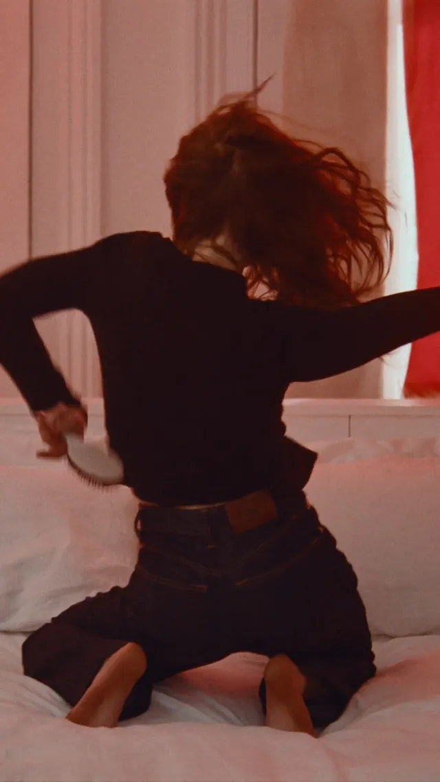 Sadie Sink kneeling barefoot on a bed in dark jeans.