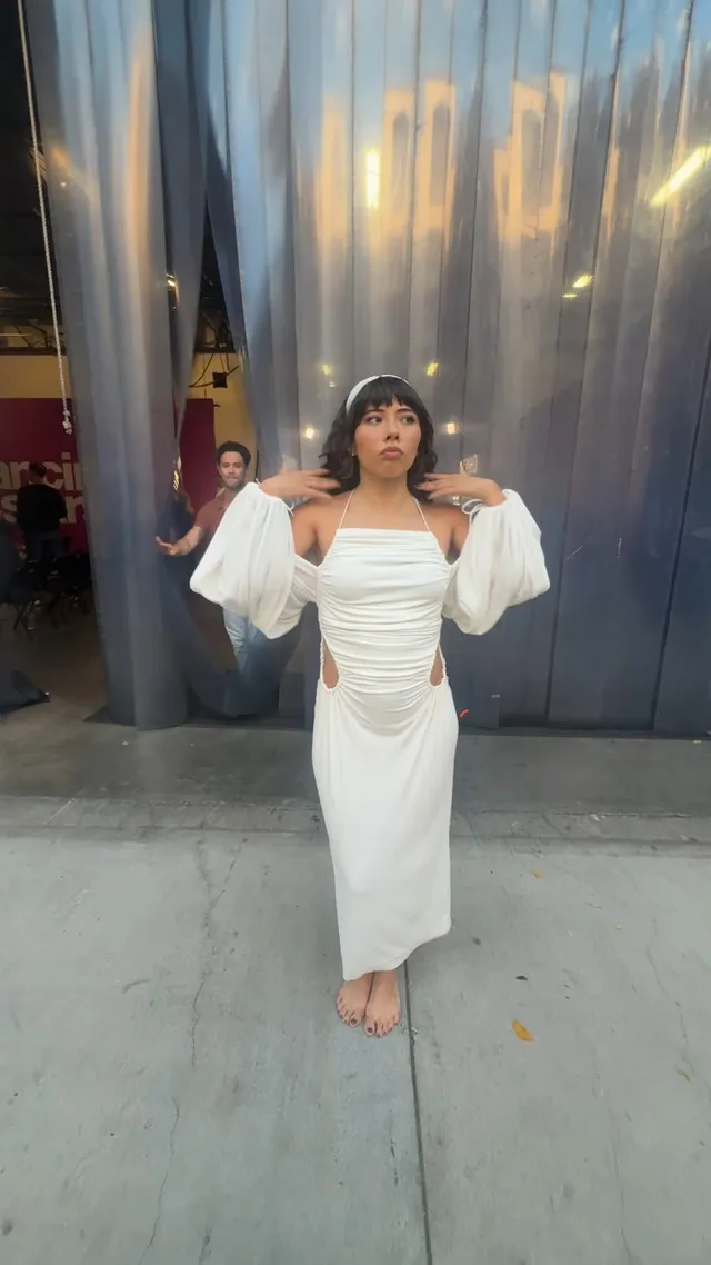 Xochitl Gomez standing barefoot outdoors in a long white dress.
