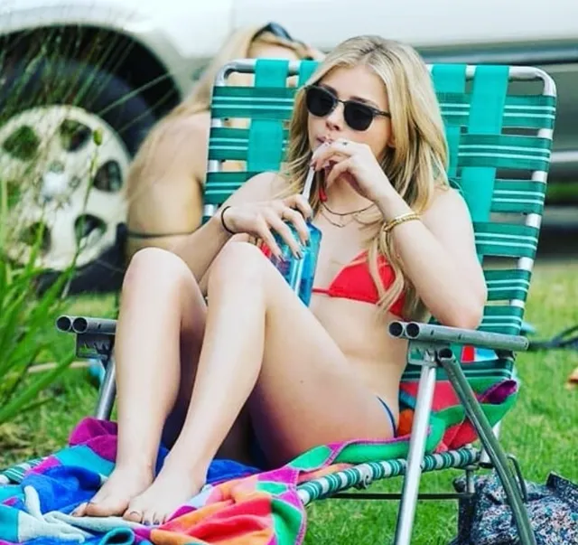 Chloë Grace Moretz sitting barefoot on a striped beach chair in a red swimsuit.