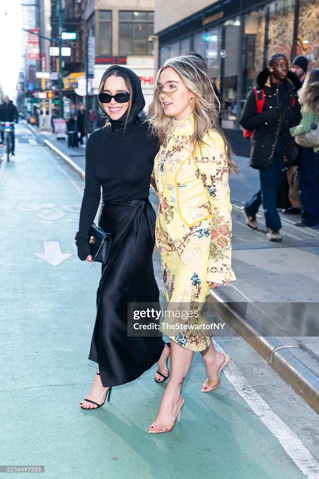 Emilia Clarke walking beside another woman in a long black dress and heels.
