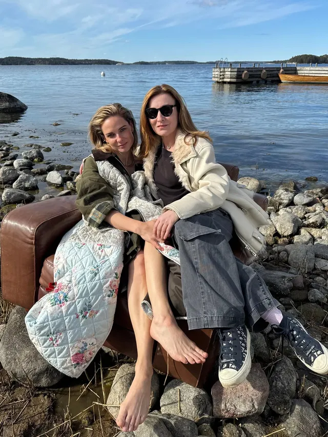 Rebecca Ferguson sitting barefoot in an armchair beside a companion on a rocky shoreline.