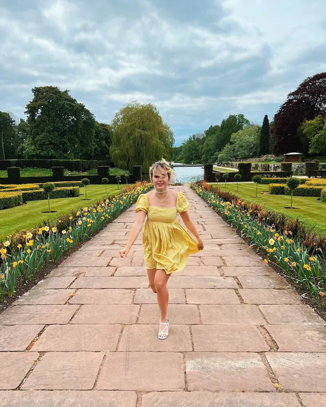 Brec Bassinger walking on a garden path in a yellow dress and white heels.