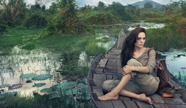 Angelina Jolie seated barefoot on a stone wall overlooking green countryside.