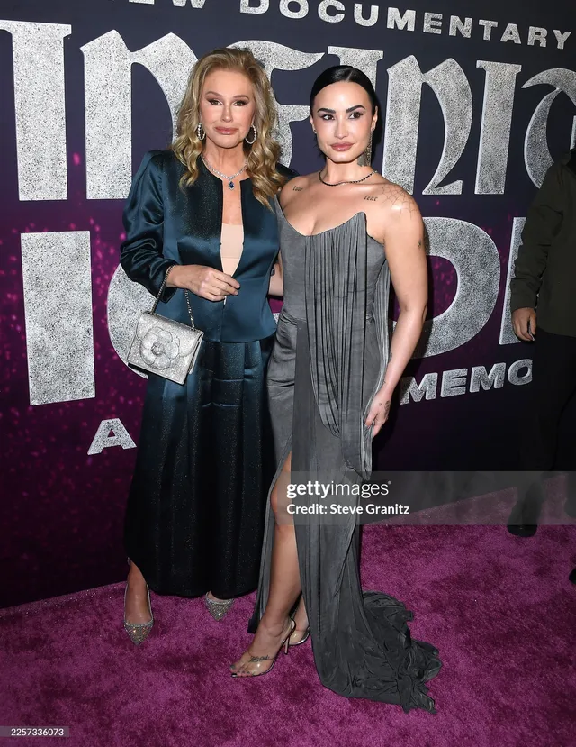 Demi Lovato posing beside another person in a grey gown and heels.