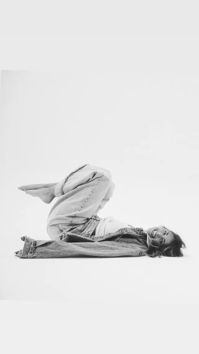 Nicole Wallace posing barefoot on a studio floor in an oversized denim outfit.