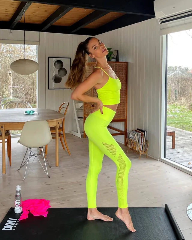 Nina Agdal standing barefoot on an exercise mat in a bright yellow outfit.